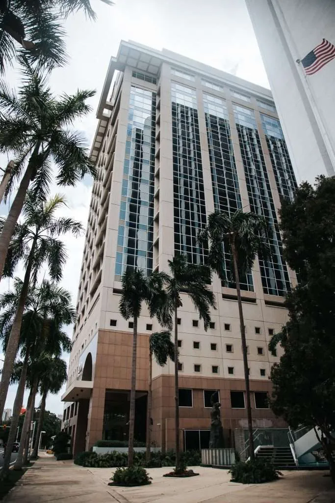 Low-angle view of a tall modern glass-office building with palm trees along the sidewalk and an American flag on the right side of the image.