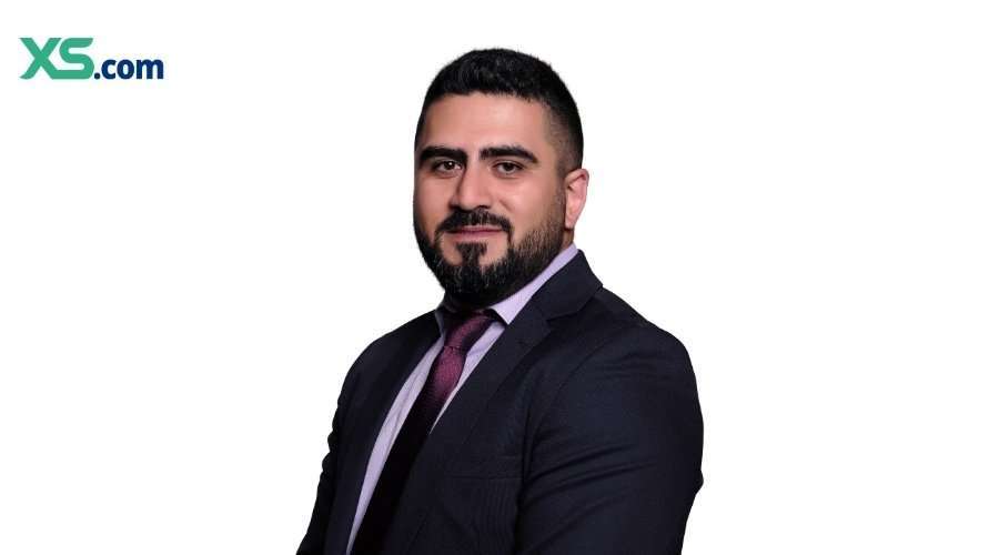 Professional head-and-shoulders portrait of a man in a dark suit and purple tie, with XS.com logo in the top-left corner on a white background, conveying a corporate profile image.