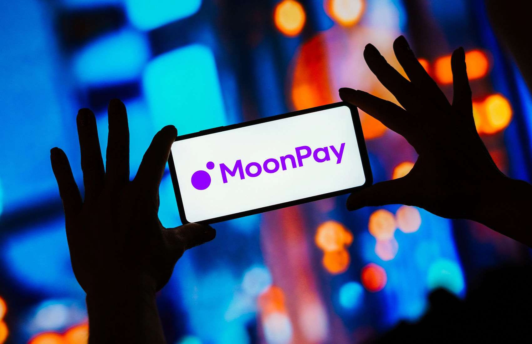 Silhouetted hands hold up a smartphone displaying the MoonPay logo against a blurred, colourful city-light background.