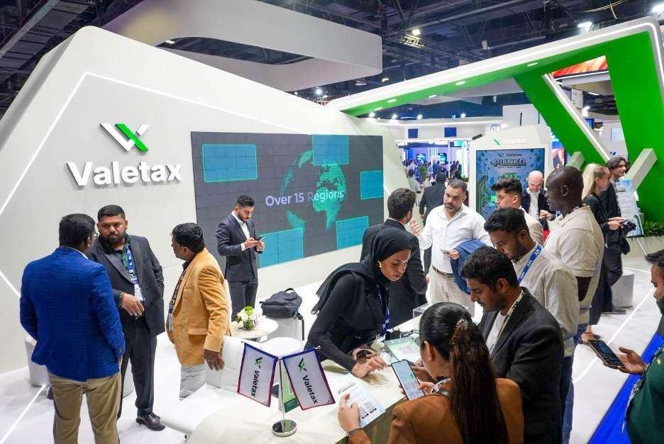 Valetax booth at a trade show with professionals gathering around a white and green display wall and a large digital screen reading over 15 regions